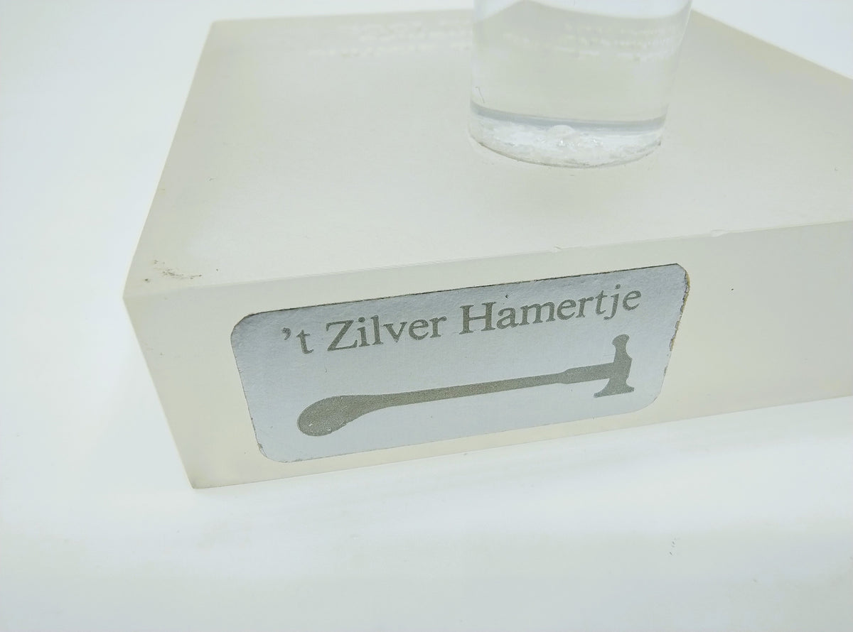 Witte sokkel met metalen label '’t Zilver Hamertje' en transparante cilinderhouder