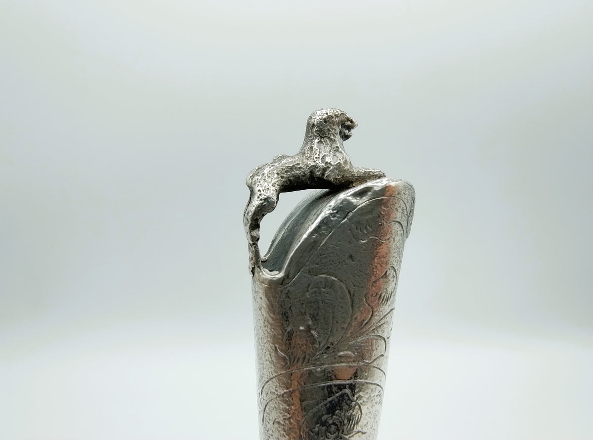 Close-up van zilveren heft met reliëf van een figuur bovenop de greep.